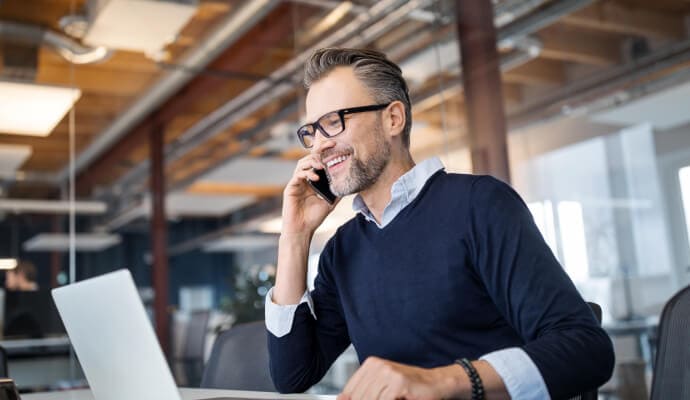 Homem sorridente com o telemóvel junto ao ouvido enquanto olha para um computador portátil