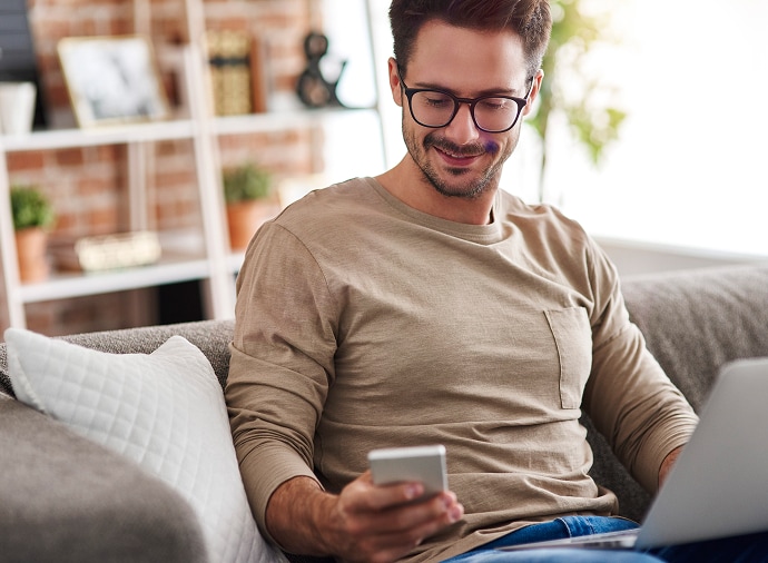 Homem sentado no sofá a olhar para o telemóvel enquanto tem um computador portátil no colo