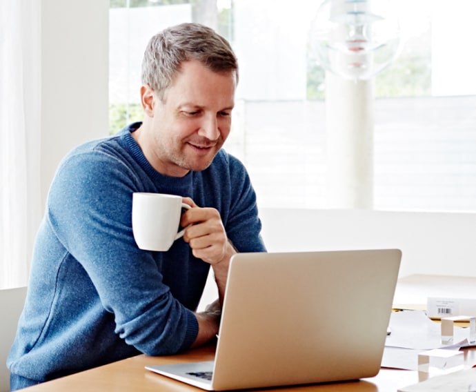 Homem a utilizar computador portátil em casa enquanto bebe café.
